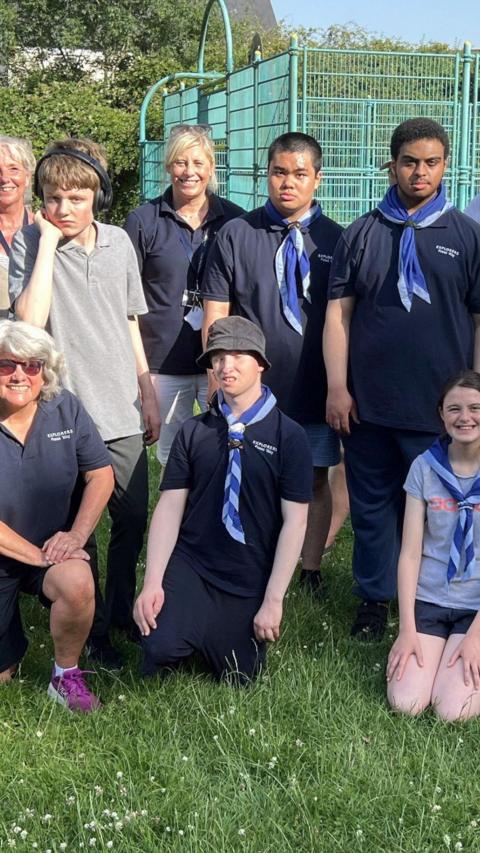 The photograph shows members of Fosseway Explorer Scout Unit and adult volunteers. Three people are kneeling in the front row (from left to right) a woman with silver hair wearing a navy scout t-shirt, sunglasses and purple trainers; a boy wearing navy scout uniform, a blue and white neckerchief and blue bucket hat; a girl with dark hair wearing a blue skirt, grey t-shirt and blue and white neckerchief. Standing in the back row (from left to right) are a woman with fair hair and patterned top; a boy with brown hair wearing dark grey trousers, a light grey t-shirt and headphones; a woman with blonde hair wearing white trousers, a navy scout t-shirt and sunglasses balanced on her head; a boy with short, dark hair wearing blue shorts, a navy scout t-shirt and blue and white neckerchief; a boy with dark hair wearing dark blue trousers, a dark blue scout t-shirt and a blue and white neckerchief.