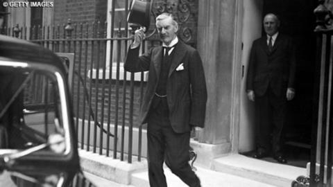 Prime Minister Neville Chamberlain doff's his hat to the camera while leaving Downing Street.