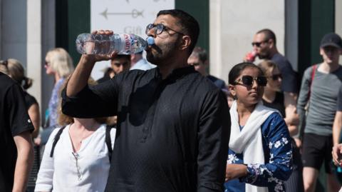 A man in a London street on a sunny day takes a drink of water from a plastic bottle. 