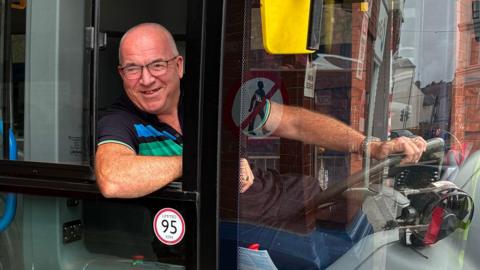 Andrew Stewart, a man in his 60s with short, grey hair and glasses, smiling. He is sitting in the driver's seat of a bus, one hand on the wheel, the other arm perched casually on the widow edge as he leans out towards the camera.