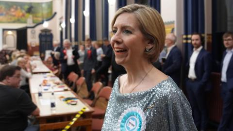 Andrea Jenkyns, who has a short blonde bob and is wearing a silver sparkly top and blue Reform badge, is smiling to the side. There is a long table behind her and several people sat down and others stood up in suits.