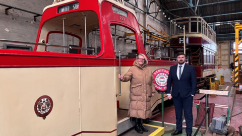 Council leader Lynn Williams, wearing a long brown winter coat, and MP Chris Webb, wearing a dark suit and red tie, stand by a red and brown tram at the museum.
