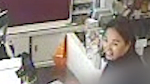 A  grainy image from CCTV of an Asian woman in dark clothing standing and at an automated till in a shop