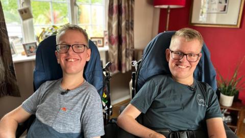 Twins Alex and Sam sat next to one another. They are sat in specialist wheelchairs. They are smiling and both have black glasses and blonde hair. 