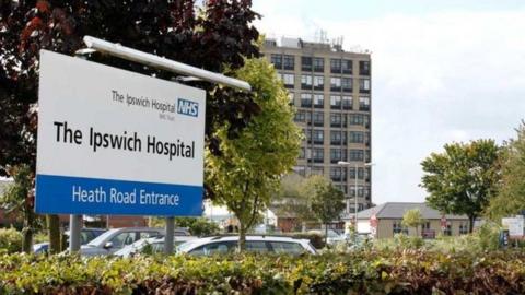 The entrance to Ipswich Hospital. A white sign details the hospital and has a blue and white NHS sign on it. Cars can be seen behind the sign parked in a car park. A multi-storey building and other smaller buildings are in the background. 