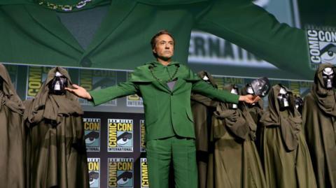 Robert Downey Jr on the Comic Con stage after revealing himself to be playing Doctor Doom. He wears a green suit and his arms are outstretched, with an iron Doctor Doom mask in one hand. He is flanked by several cloaked Doctor Doom lookalikes.
