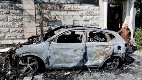 A Palestinian man walks past a torched car in the village of Jit, in the north of the occupied West Bank (16 August 2024)