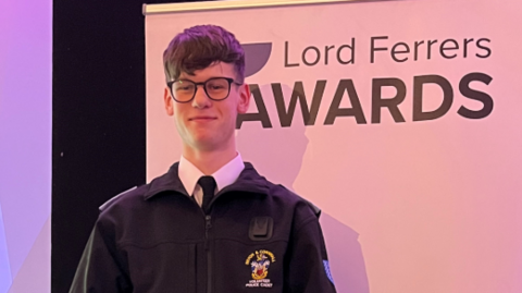 Caolan Hutchins in his black cadet uniform. He has black glasses and hair and is standing next to the award banner