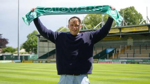 Yeovil goalkeeper stands on the pitch at Huish Park holding a scarf saying glovers, to celebrate signing a contract at the club