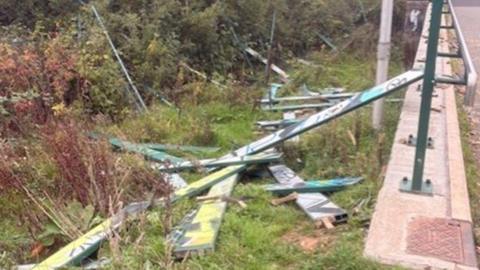 Fencing from a skate park in Exeter which has been left strewn on the ground after being vandalised. The green panels have graffiti on the them. Bushes are next to the discarded fence panels.