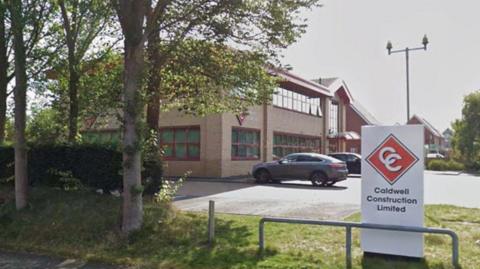 A white totem on a patch of grass alongside a car park with a building and parked cars in the background. There are trees along the verge in the foreground to the left hand side of the image.