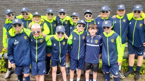 A team of young footballers in team colours of blue and yellow, pose in two rows.