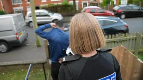 A woman with blonde short hair in a police vest. The officers have arrested a man in a blue shirt. They are walking down steps from a house, onto a street with parked cars