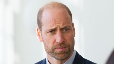 Prince William looks into the distance at an event while wearing a dark blazer and a light blue shirt. He has a short beard covering his lower face. He is stood in front of a white wall.