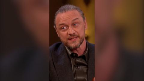 James McAvoy has greying hair tied back, and a beard and moustache. He is wearing a shiny black shirt and a black jacket.