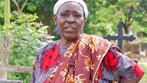 Pudensiana Chumbi, wearing a headwrap and dressed in a flower print dress with long scarf draped over one shoulder, a stands in the graveyard in Morogoro with a hand on her hip looking at the camera.