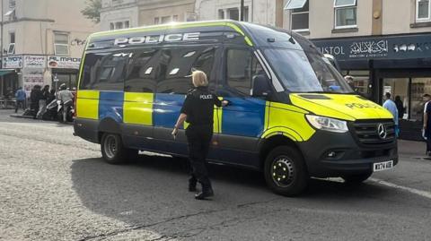A police van on a closed road. A police officer stands outside it.