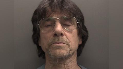 Police mugshot of defendant with eyes closed, shoulder length dark hair, glasses and a goatee 