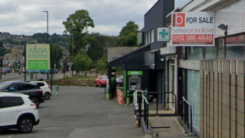 A strip of shops on a main road. In the foreground is a For Sale sign on an empty building. Behind is a pharmacy and an Asda.