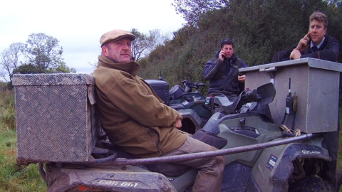 A man sat on a quad bike with two men using their mobile phones behind him