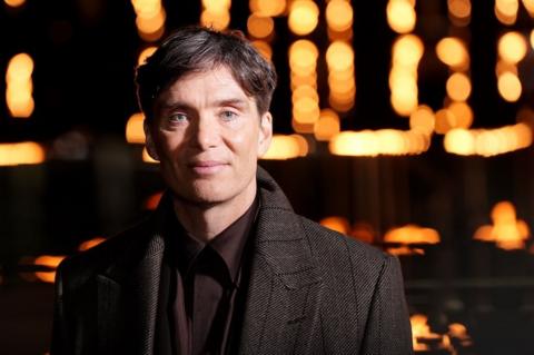 Actor Cillian Murphy stares into camera with a gentle smile on his face. He's wearing a brown coat and burgundy silk shirt.
