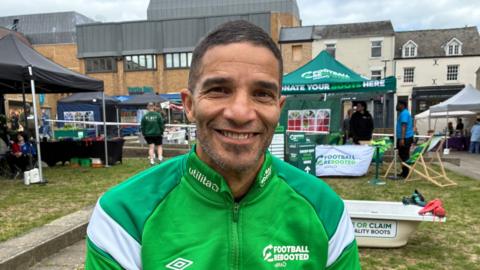 David James in Peterborough. He is wearing a green and white fleece. He has short dark hair and a very short beard and is smiling broadly. Behind him are various gazebos with people sitting or standing underneath them. Wording on a gazebo says "DONATE YOUR BOOTS HERE"