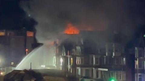 Water from a fire engine's hose can be seen spraying to a building on fire in Newquay. There are orange flames at the top of the building. It is a low-quality image taken at night.