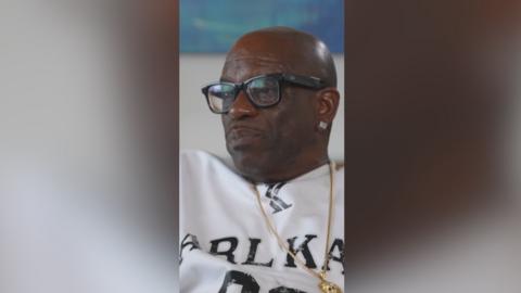 A man with a gold chain and black glasses speaks into the camera.