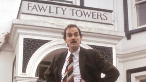 A man with a moustache wearing a suit in front of a hotel saying Fawlty Towers
