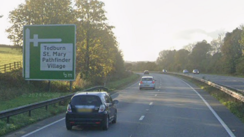 Cars travelling along a dual carriageway. A large green sign shows directions towards Tedburn St. Mary and Pathfinder Village. Trees are on the edge of the carriageway. A central reservation seperates the westbound carriageway from the eastbound carriage.