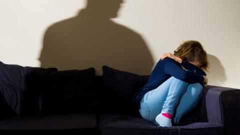 A woman cowering in the corner of a dark sofa. She is wearing jeans and a dfark blue top and her head is in her hands, with her knees tucked up to her chin. There is a shadow of a man above her. 