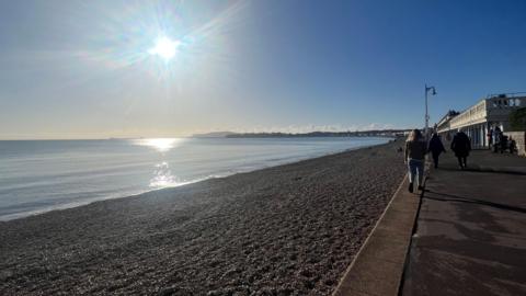 blue sea with stoney beach next to a concrete esplanade. People are walking alongside the beach. A low sun shines in a cloudless sky.