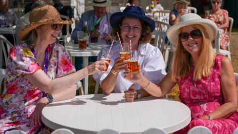 Three women sit down enjoying glasses of drinks at the Taunton Flower show. They are sitting under a sunshade and are all wearing sun hats and are around a white table