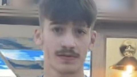 Taylor Charlton is wearing a blue shirt with a white Nike logo on it. He has a short moustache and short hair.