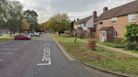 Lancaster Square in Lyneham. There are rows of parking spaces to the left, with residential properties to the right and a grassy verge.