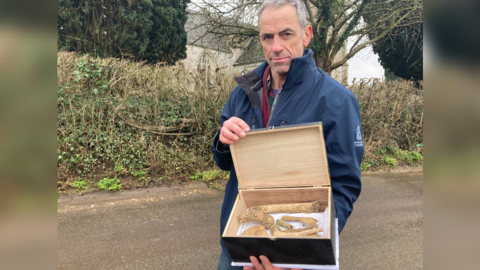 A man wearing a blue coat looking at the camera. He's holding a box open which contains a selection of bones 