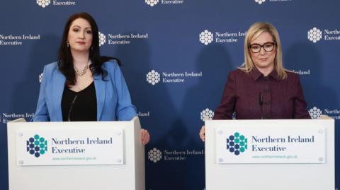 Emma Little-Pengelly and Michelle O'Neill standing at podiums with the logo of the Northern Ireland Executive on them. They are standing in front of a background with the same logo