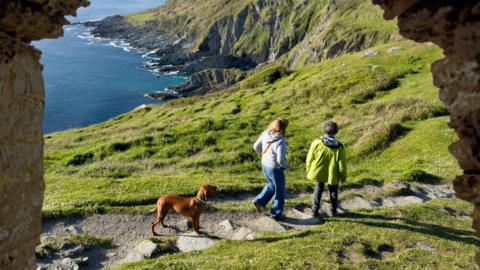 Two people are walking along a grassy path accompanied by a brown dog. The path winds toward dramatic, rocky cliffs that drop into the sea.