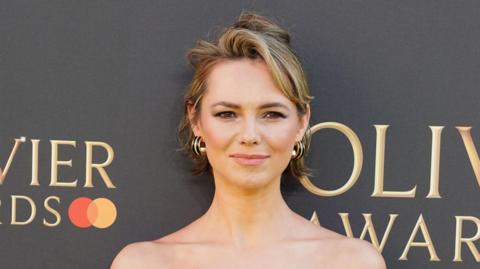 Head and shoulders of Kara Tointon. She has dark blonde hair in a messy ponytail with black and gold hoop earrings. She is standing in front of a grey background that says Oliver Awards in gold writing. 