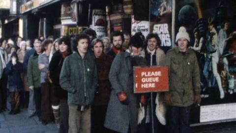 A queue of people, all wearing warm coats, standing in front of Star Wars posters outside a cinema.