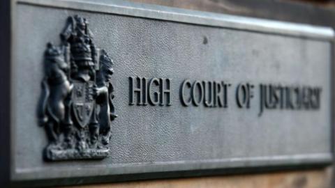 Grey sign saying 'High Court of Justiciary' outside the High Court in Edinburgh.