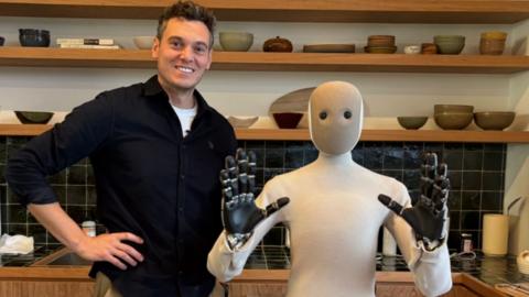 A man, Joe Tidy, wears a black sweater while standing next to a humanoid robot