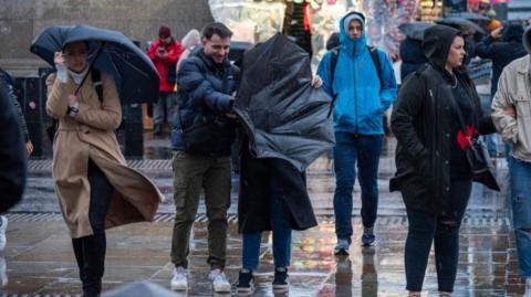 A couple struggle to keep their umbrella up, and a man behind walks with his coat zipped up to his face, as cold weather continues in the UK.