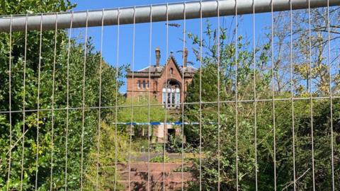 An old sandstone building viewed through fencing with police tape in front of it and some overgrown bushes