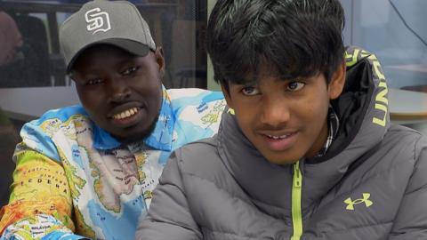 A man and a boy sat smiling, one in a grey cap and a shirt with a world-map design on it, and the other in a grey and green coat.