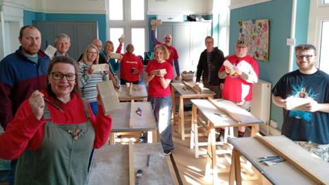 A group of about dozen men and women make blocks for the monument in a workshop. Brushes and carving tools are scattered on the wooden tables. The people are holding up their cobbles as sunlight streams in through the workshop windows. 