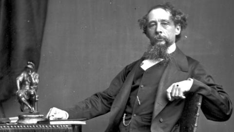 Charles Dickens (1812-1870), English novelist and one of the most popular writers in the history of literature, sitting at a writing desk, with a small statue on the desk
