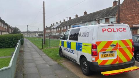 A Cleveland Police van is parked between two rows of terraced housing with a grass area in between. Police tape can be seen cordoning off the scene.