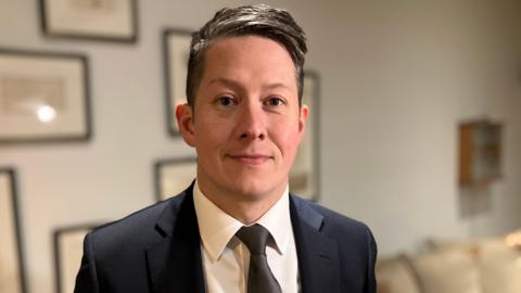 A man in a black suit jacket, black tie and white shirt. He has dark hair, combed to the side. He is smiling at the camera, standing in a room with cream sofas and black framed prints on grey walls.