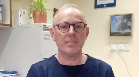 Man sitting as a desk with a computer and some papers on it. He is wearing medical scrubs.